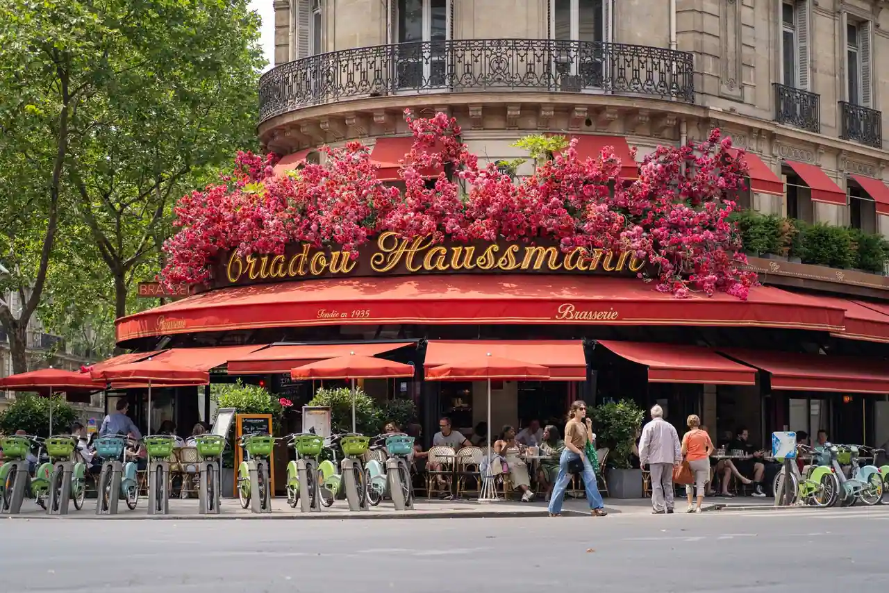 Façade d'angle d'une brasserie parisienne aux stores rouges, fleurie, avec sa terrasse animée