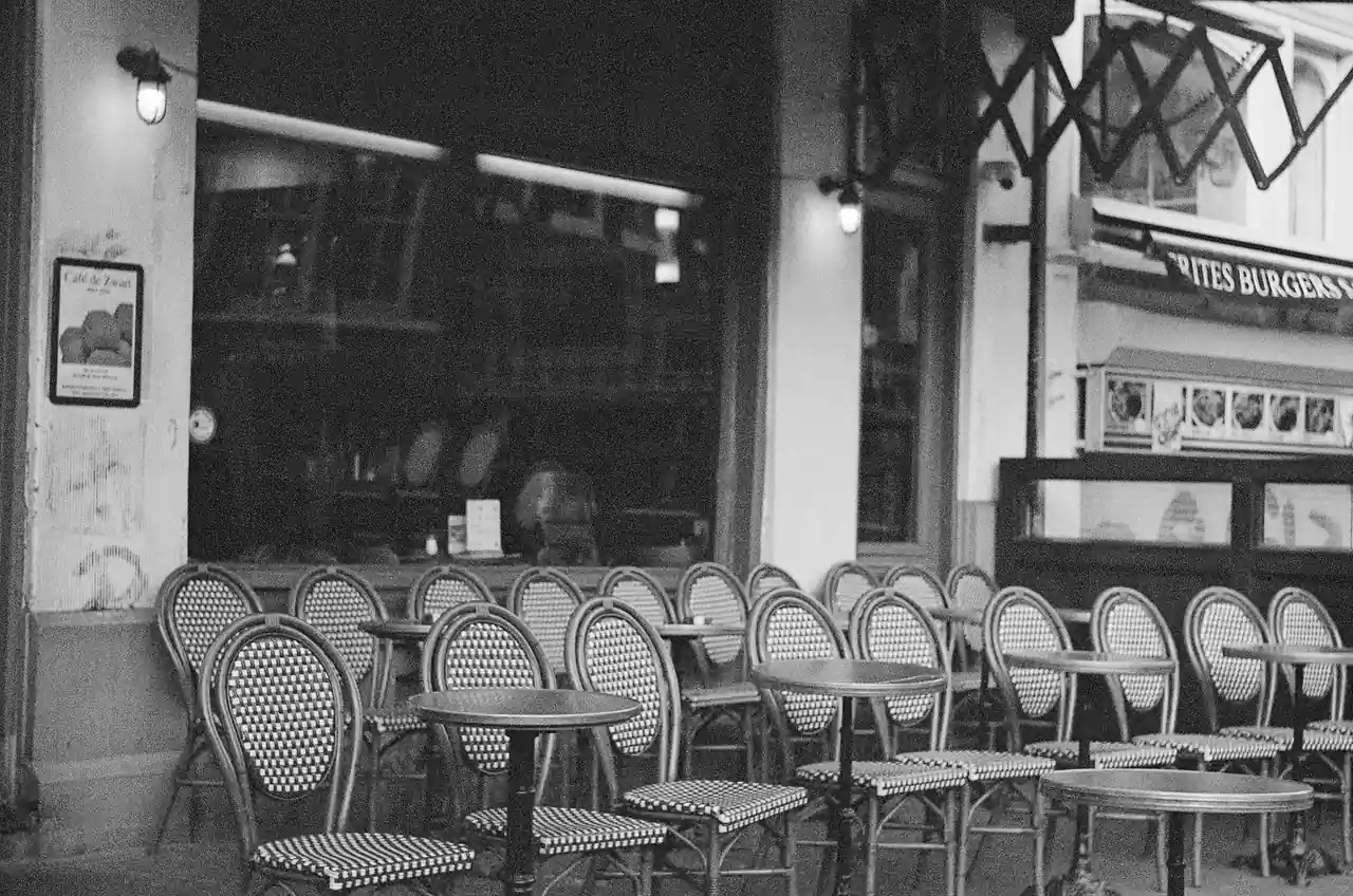 Tables rondes et chaises en rotin alignées à la terrasse d'un café parisien, en noir et blanc.