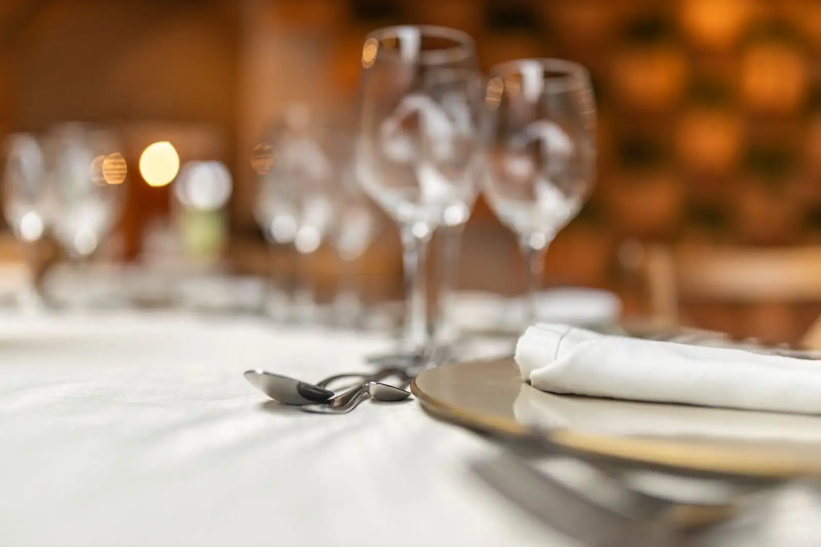 Table de restaurant dressée : assiette avec serviette pliée, couverts et verres à vin sur une nappe blanche.