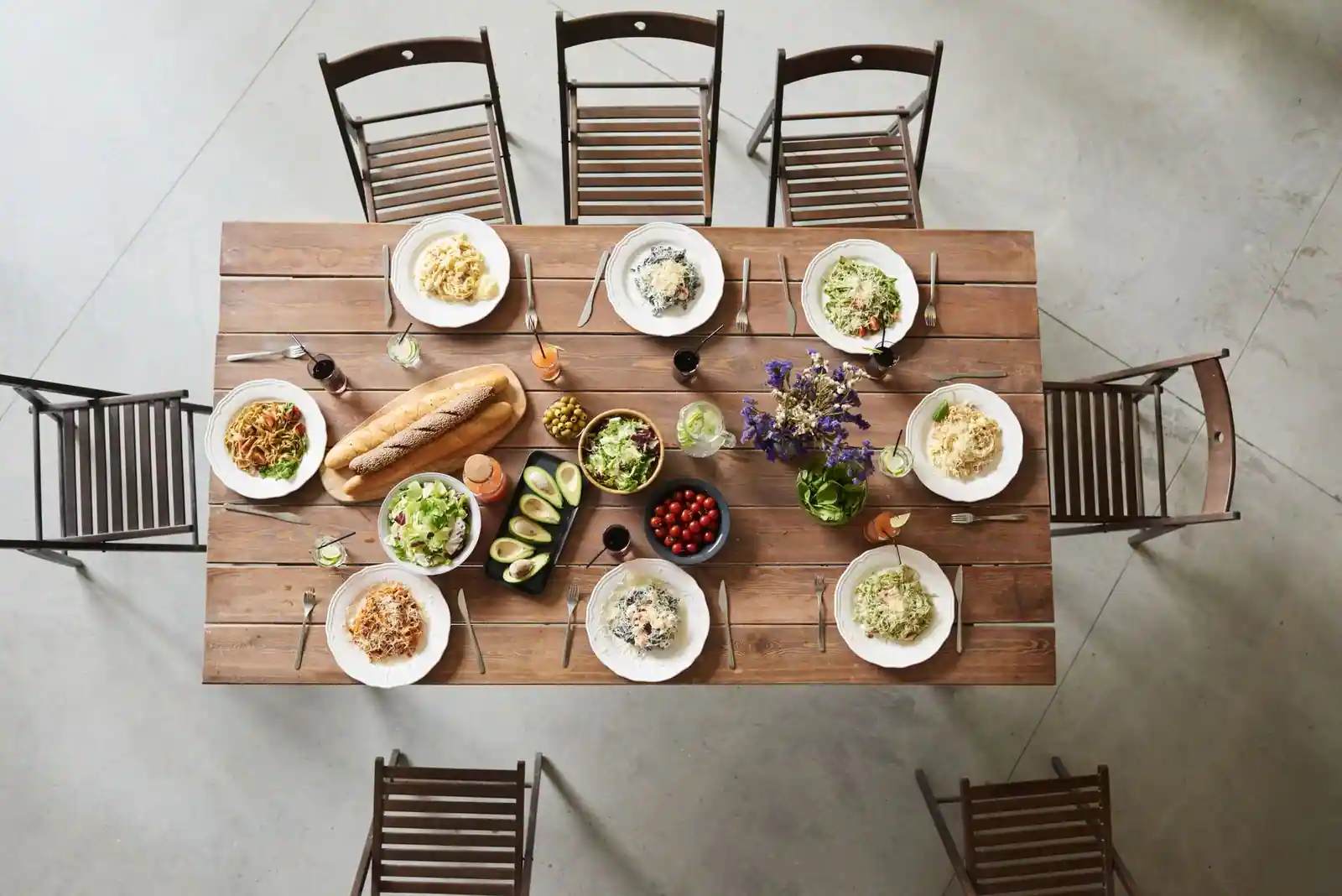 Table en bois vue de dessus, dressée pour un repas convivial avec plusieurs plats de pâtes, salade et pain