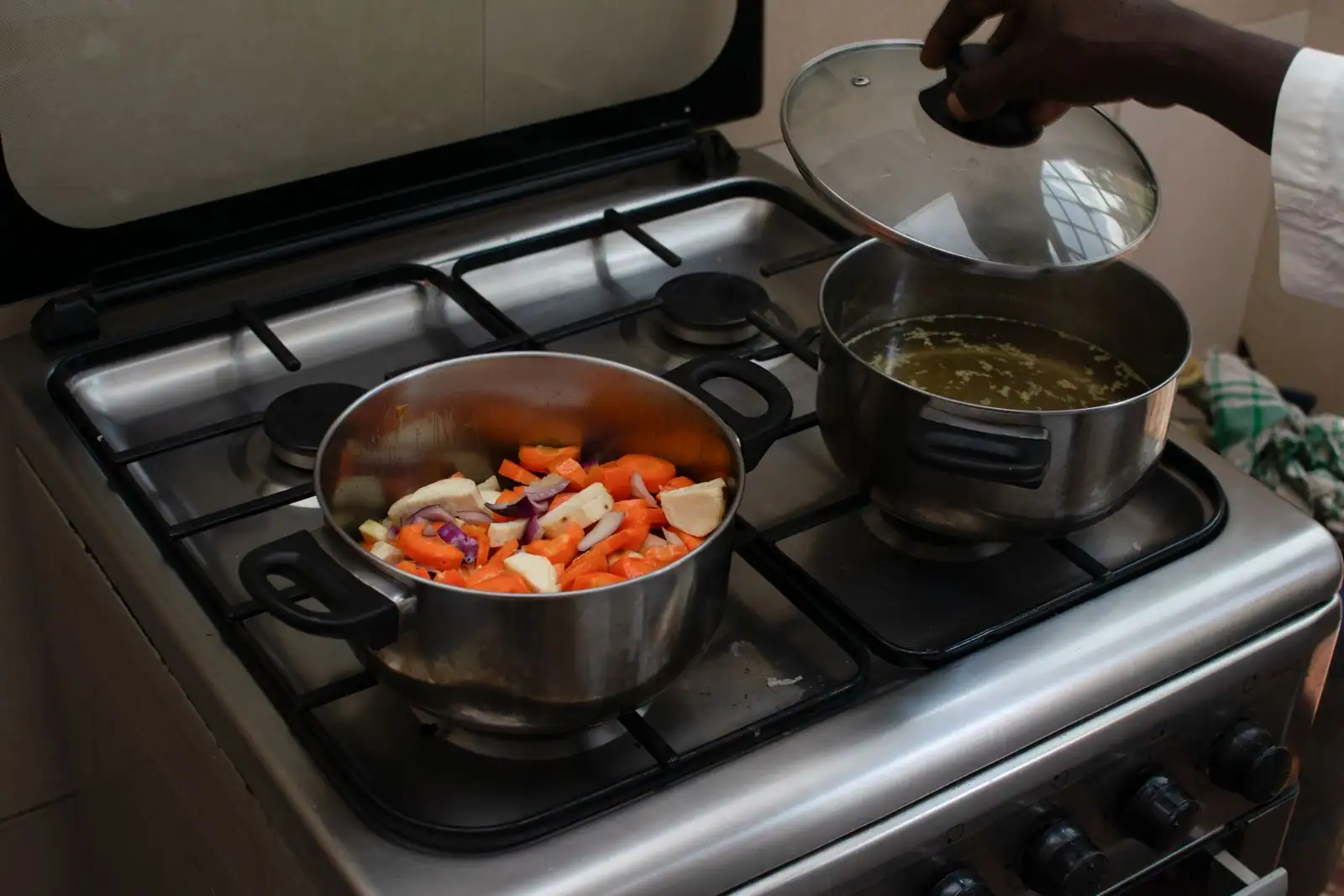 Légumes coupés qui mijotent dans une casserole sur une gazinière, une main soulevant le couvercle d'un autre faitout.