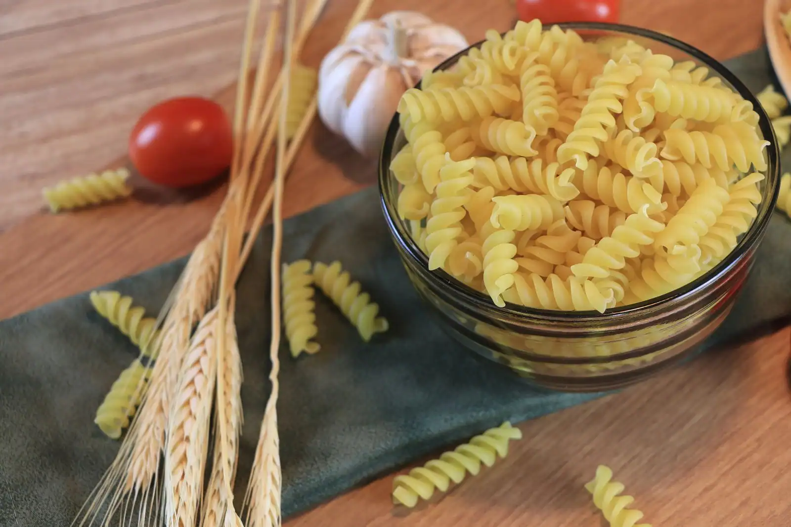 Bol de pâtes sèches sur une table en bois, entouré d'épis de blé, d'ail et d'une tomate cerise.