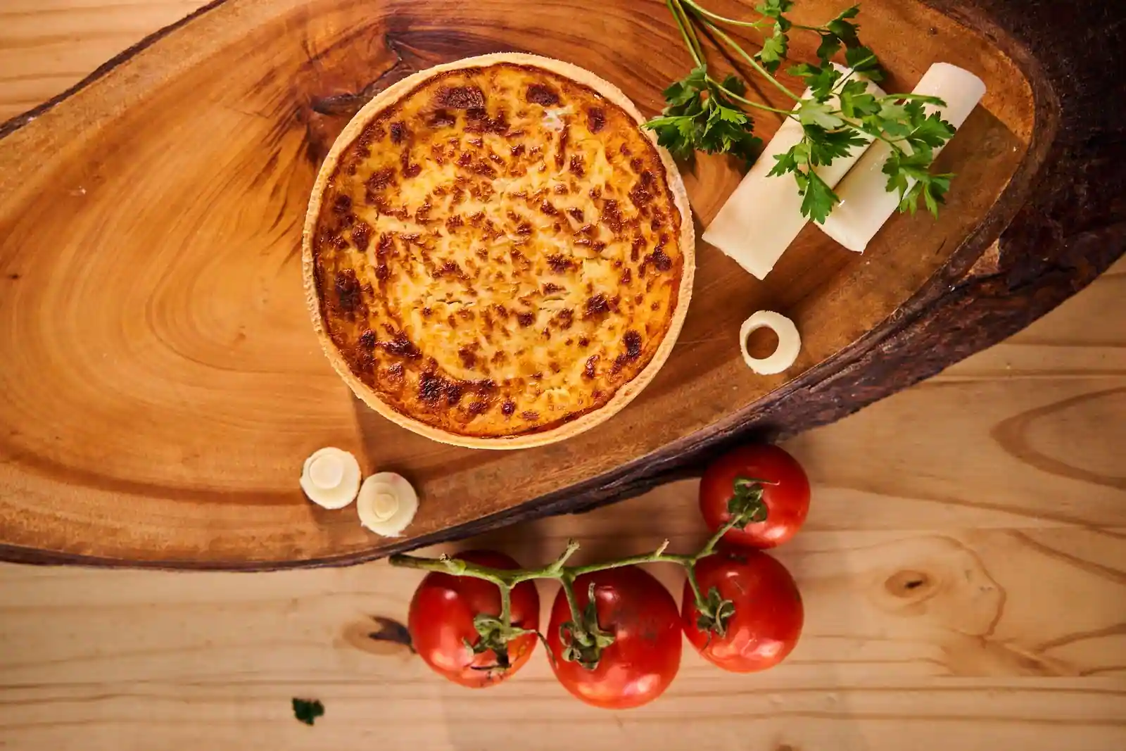 Quiche entière à la croûte dorée sur une planche en bois, entourée de tomates et de persil.