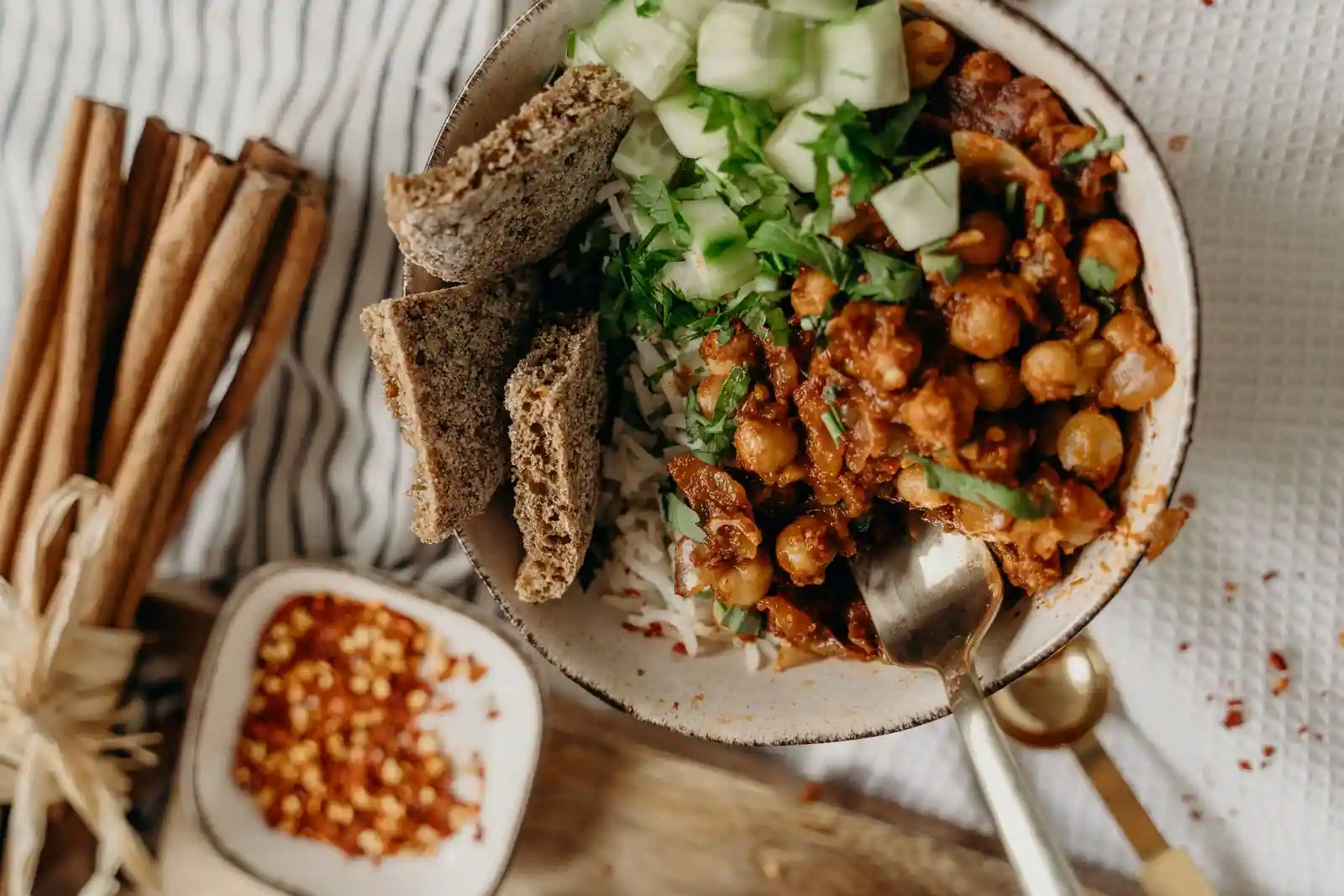Bol de pois chiches en sauce épicée sur du riz, garni de concombre et de coriandre, avec du pain à côté.