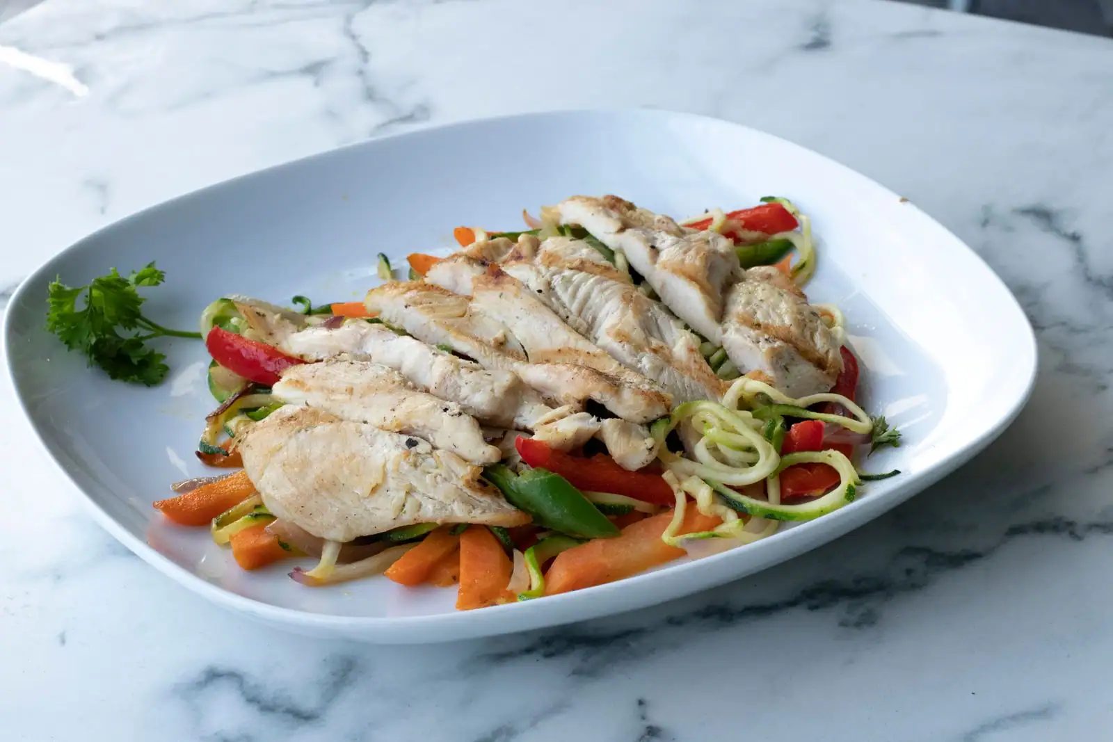 Blanc de poulet grillé tranché sur des tagliatelles de courgette, carottes et poivron rouge, dans une assiette blanche.