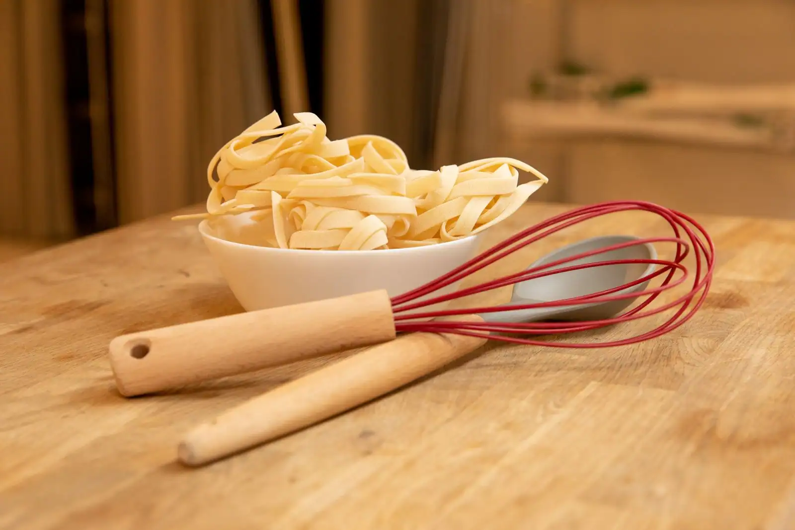Bol de tagliatelles fraîches, un fouet rouge et un ustensile en bois sur une table en bois