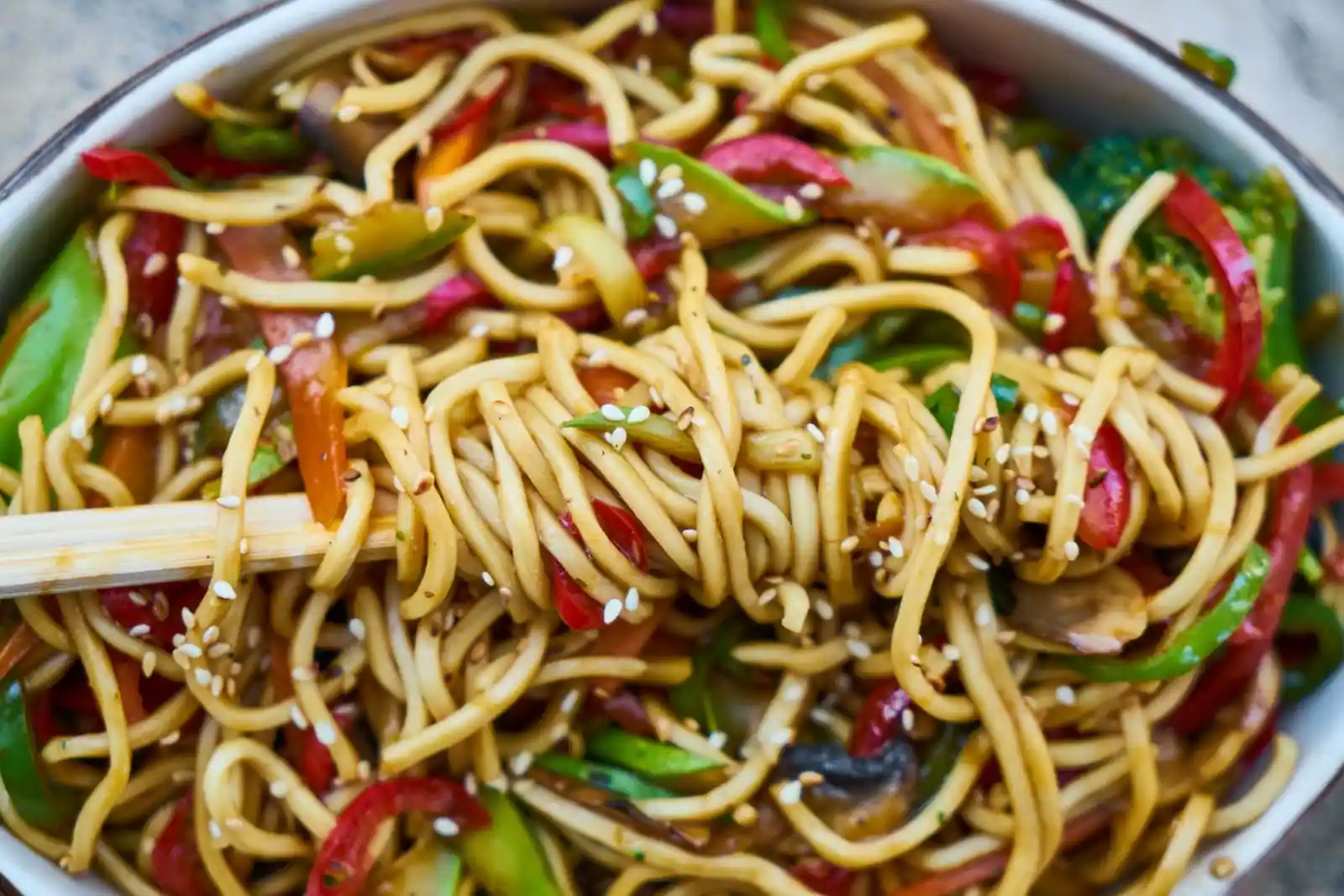 Nouilles sautées au wok avec poivrons et légumes, parsemées de sésame, servies dans un bol.