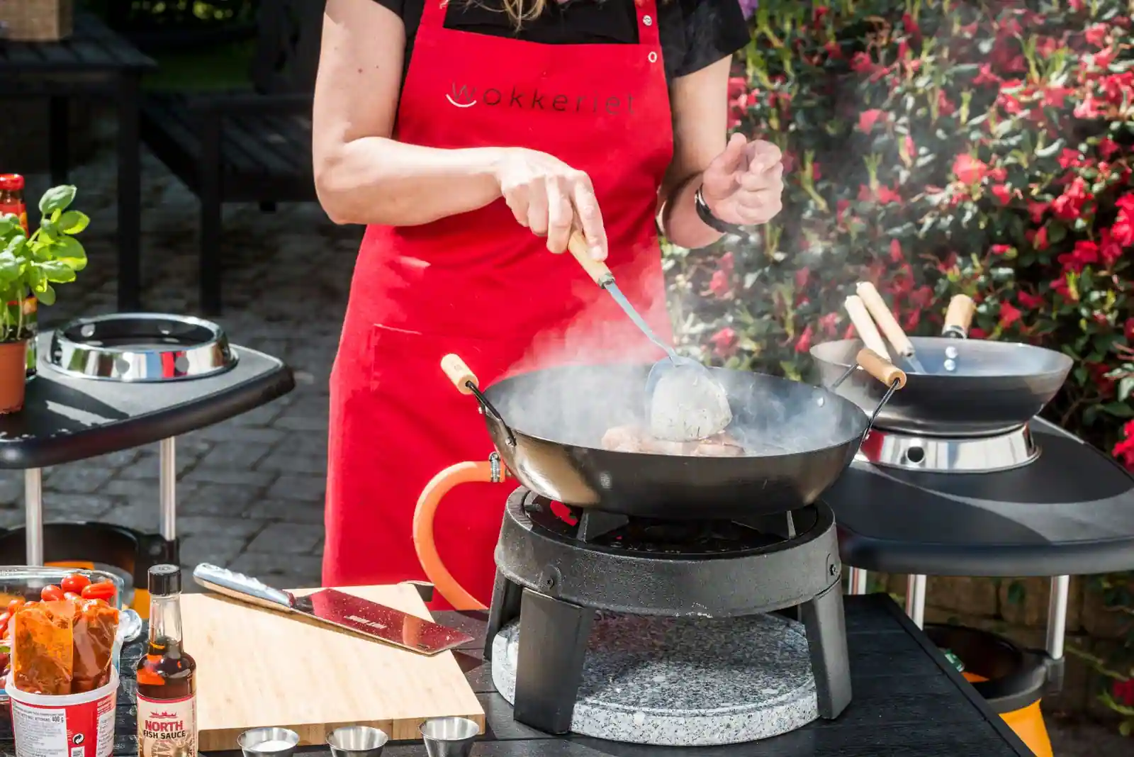 Personne cuisinant au wok en plein air sur un réchaud, vapeur s'élevant et ingrédients préparés à côté.