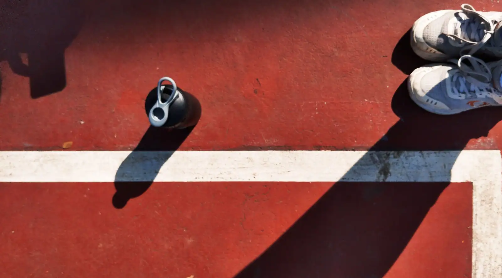Gourde de sport posée sur un court en résine rouge, près d'une paire de chaussures.