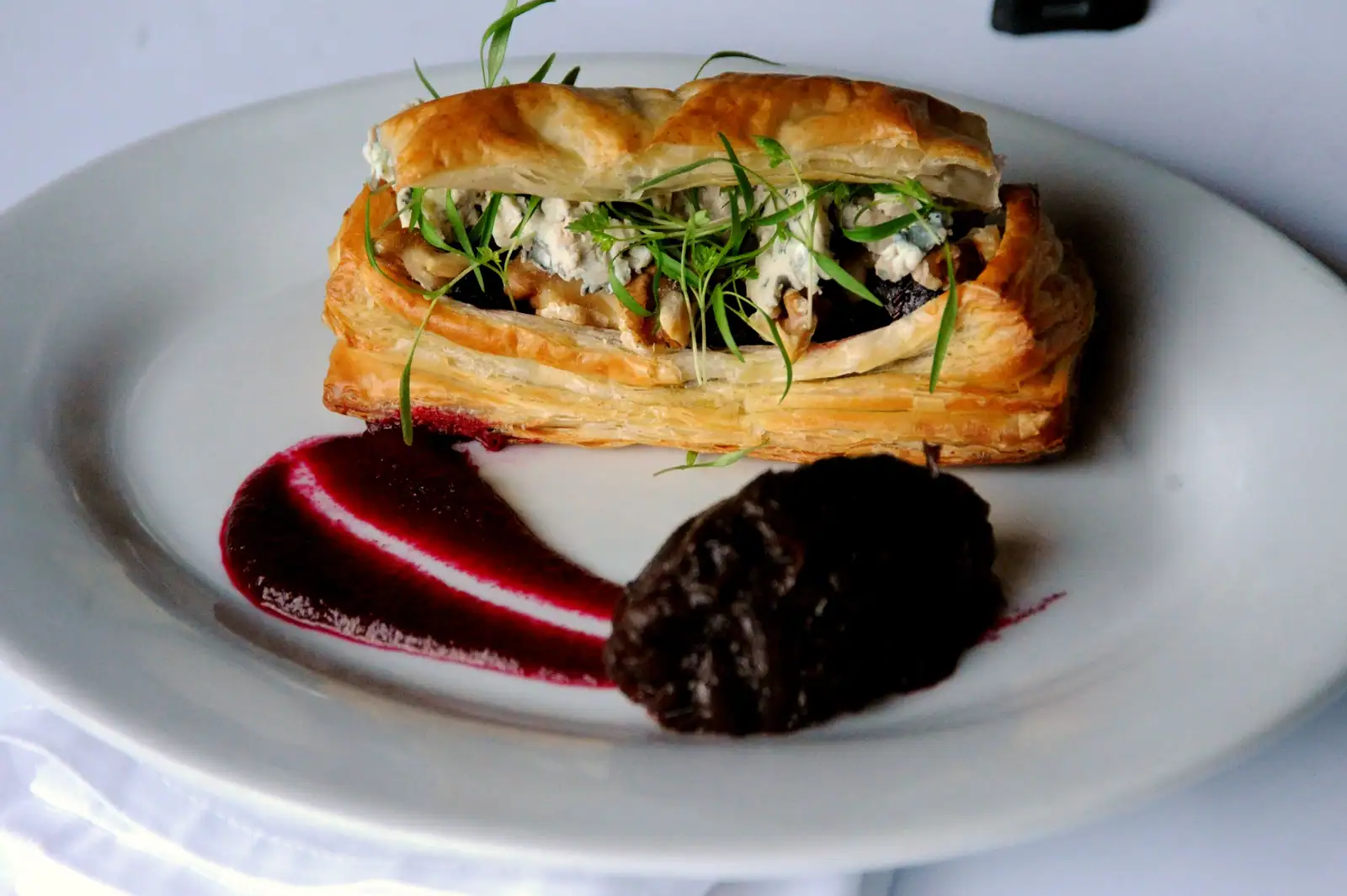 Feuilleté salé ouvert, garni de champignons et de fromage, servi avec un coulis sombre sur assiette blanche