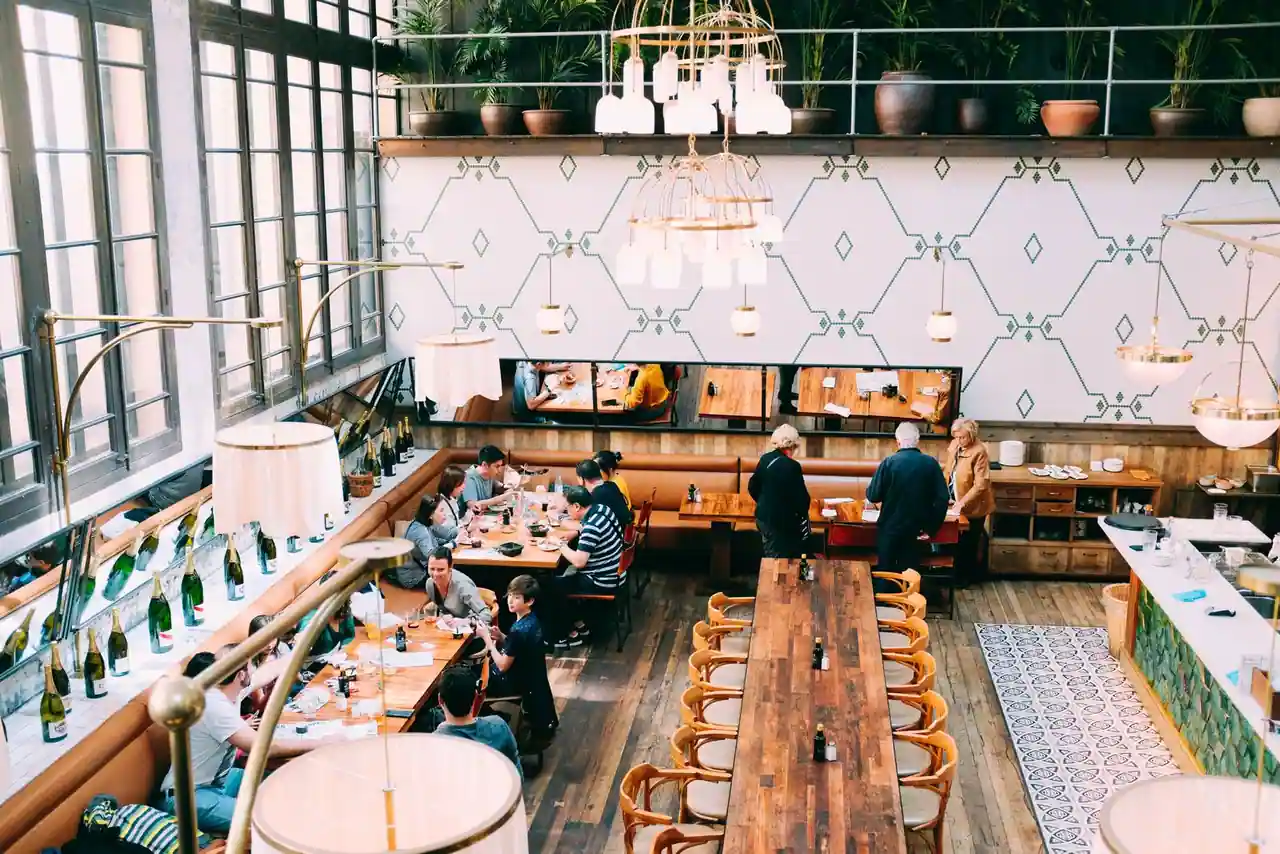 Grande salle de restaurant vue depuis un niveau supérieur : longues tables, banquettes et hautes fenêtres