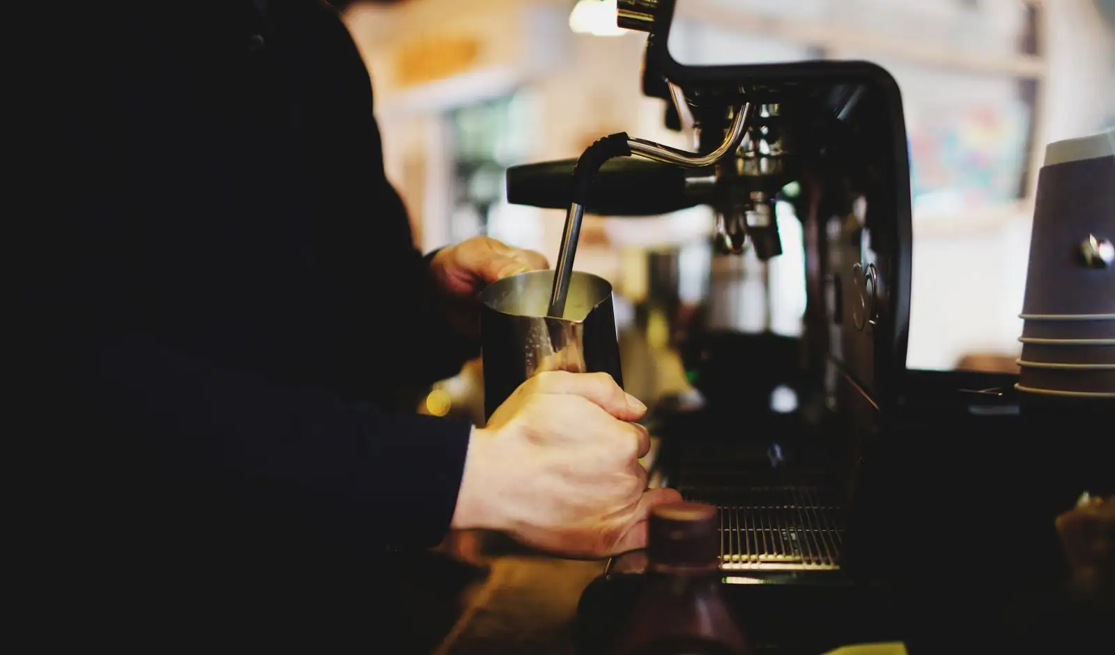 Barista faisant mousser du lait à la buse vapeur d'une machine à espresso, derrière un comptoir.