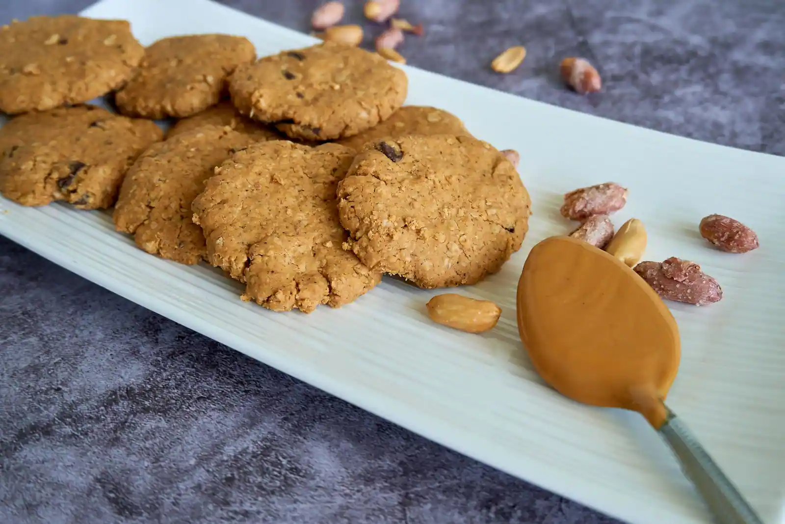 Cookies au beurre de cacahuète sur un plat allongé, avec une cuillère de purée d'arachide et des cacahuètes.