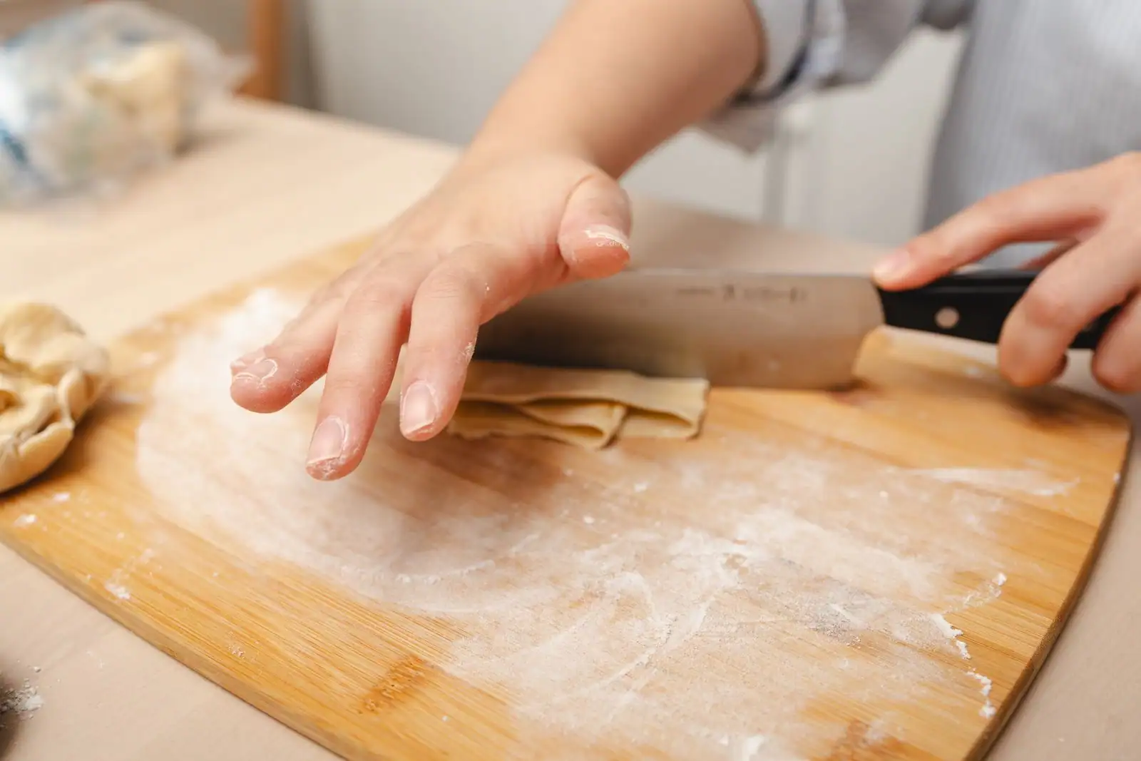 Mains découpant au couteau une feuille de pâte fraîche pliée sur une planche en bois farinée