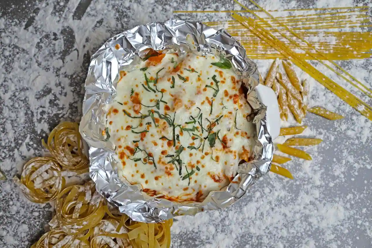 Gratin de pâtes gratiné au fromage fondu et parsemé de basilic, entouré de pâtes crues et de farine.