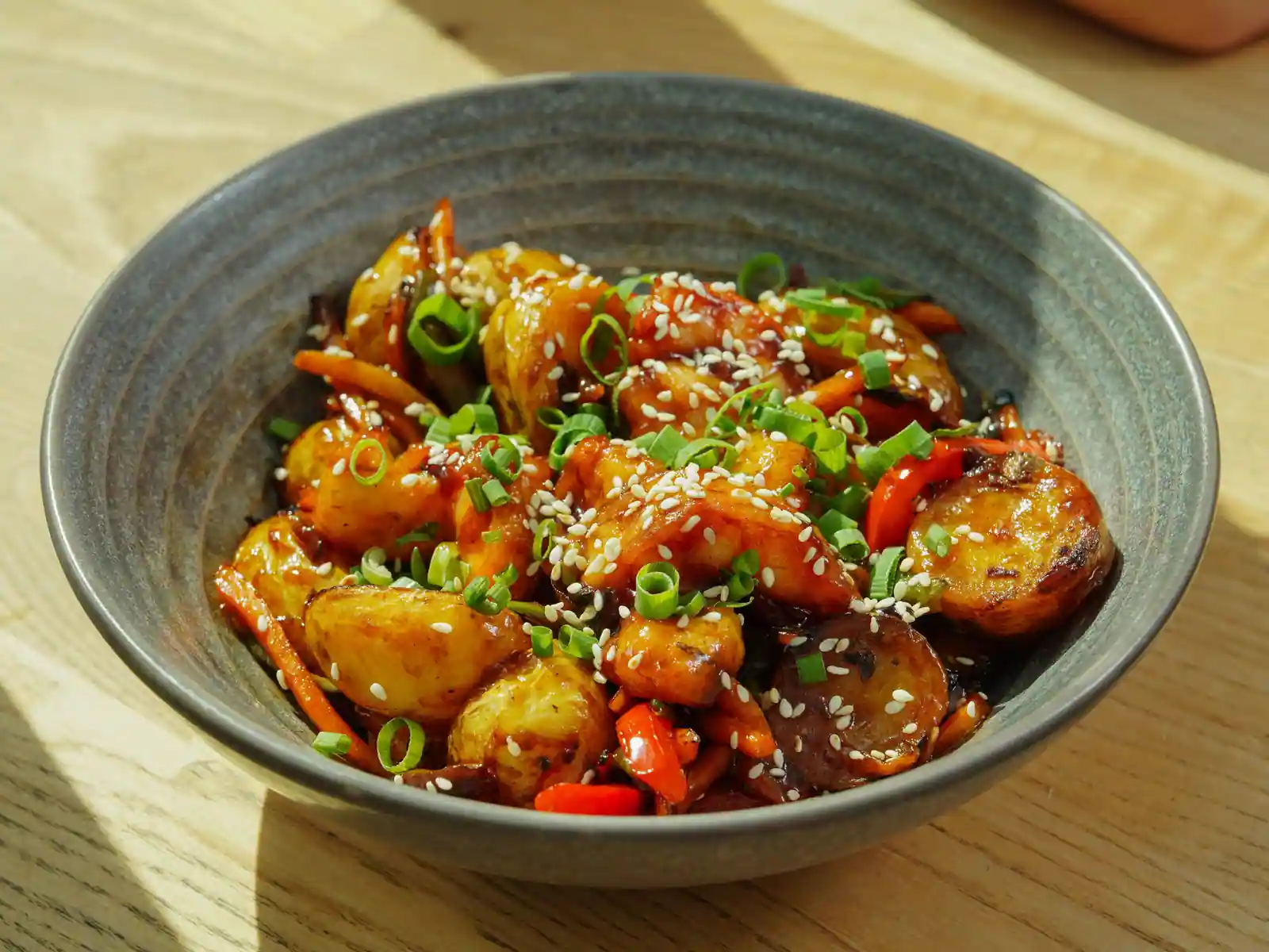 Bol de légumes rôtis nappés de sauce, parsemés de graines de sésame et d’oignon vert, sur une table en bois.