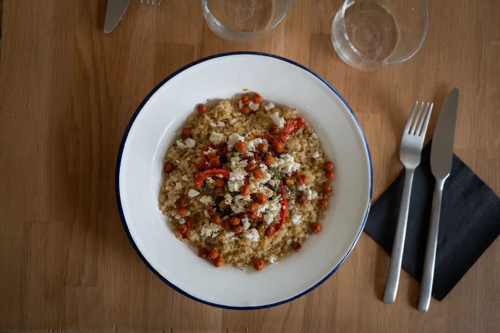 Assiette de quinoa aux pois chiches rôtis, lanières de poivron rouge et fromage blanc émietté, sur une table en bois.