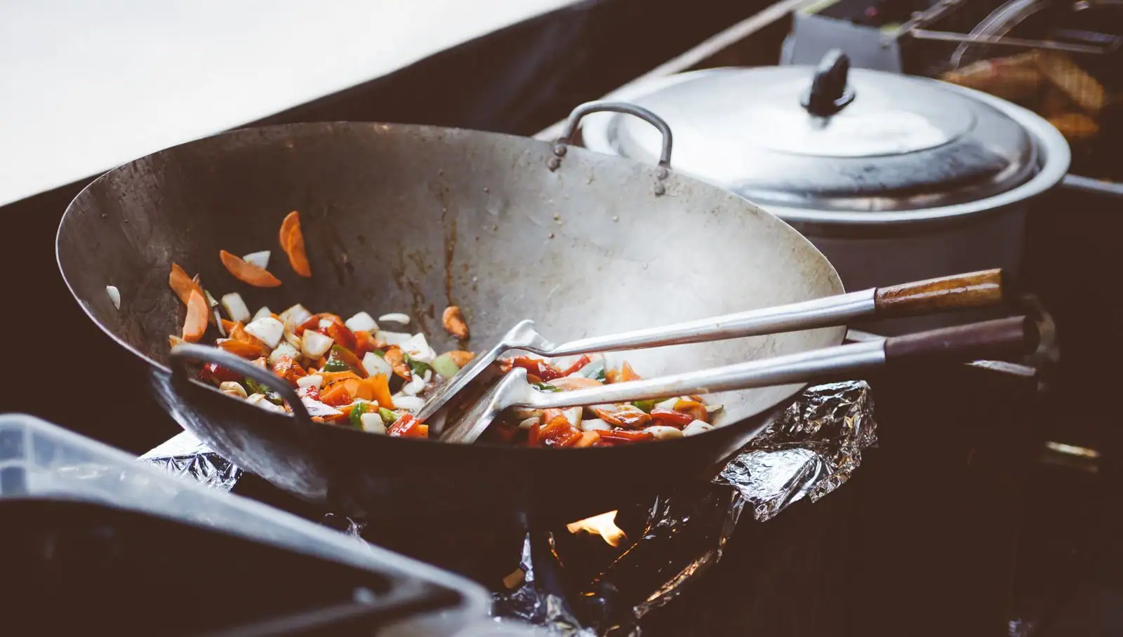 Grand wok en acier dans lequel sautent des légumes coupés, avec ses spatules posées sur le bord.