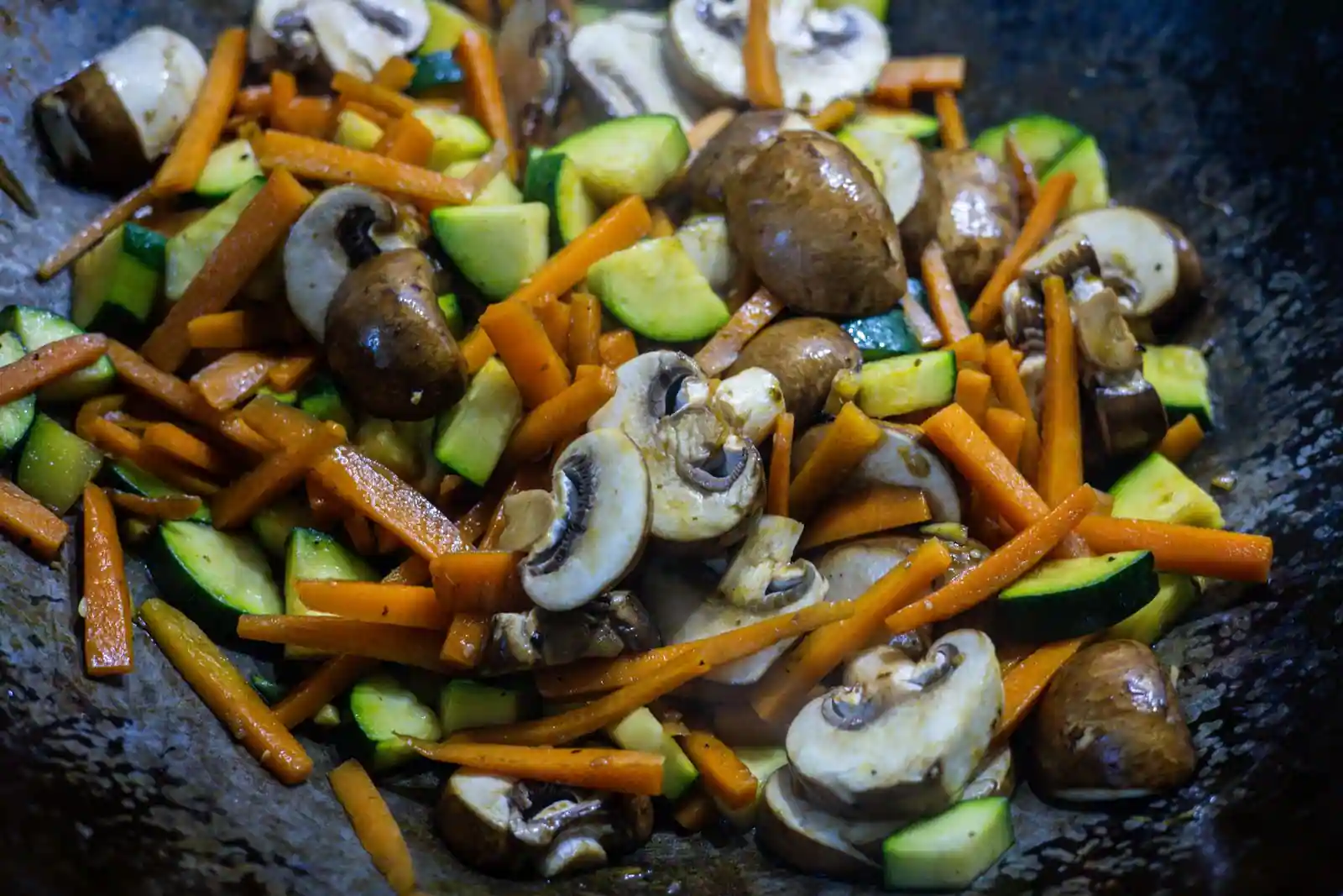 Carottes en bâtonnets, courgettes en dés et champignons sautés à feu vif dans un wok en acier noirci
