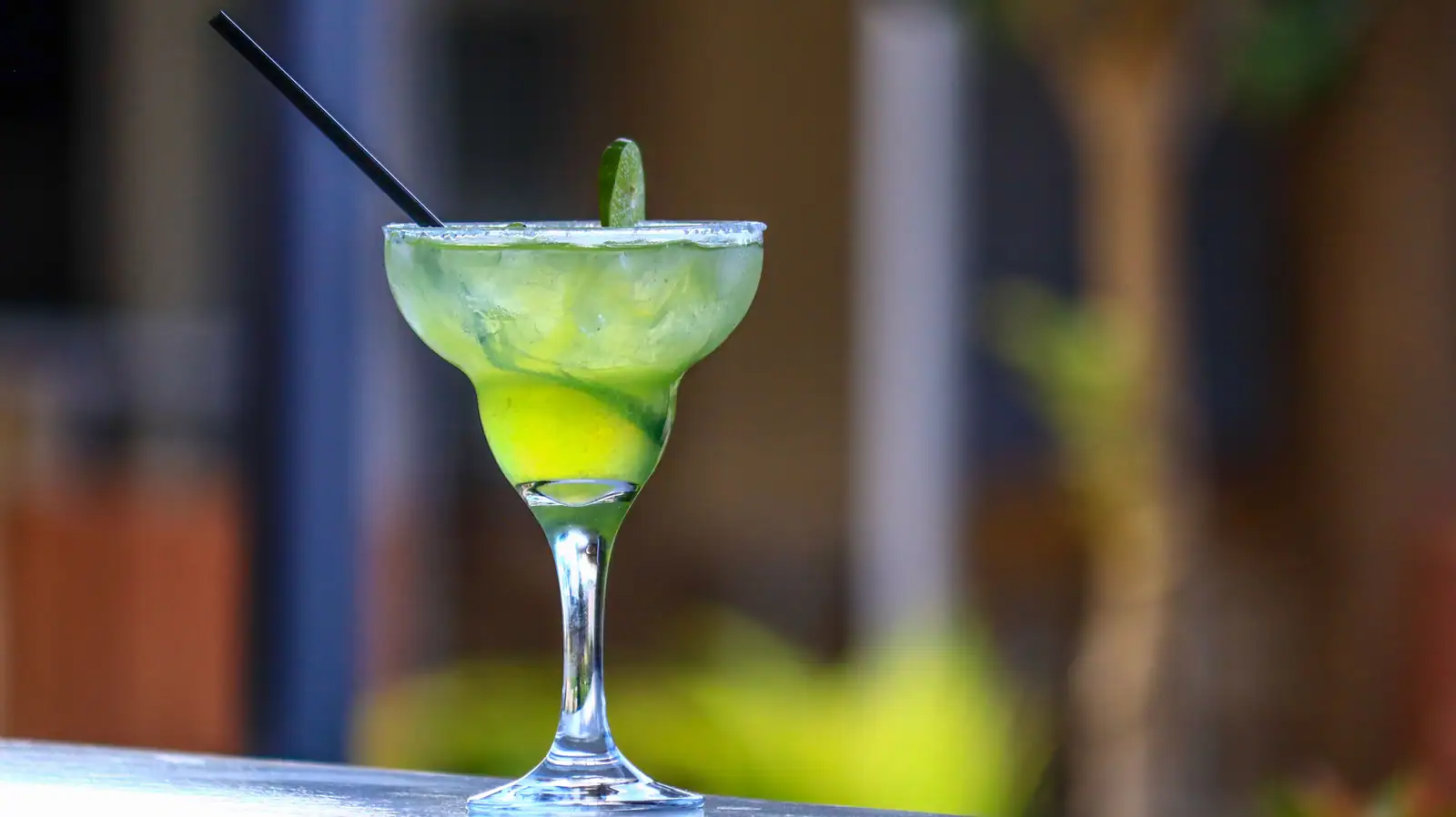 Margarita servie dans un verre à bord large et salé, avec un quartier de citron vert et une paille noire.