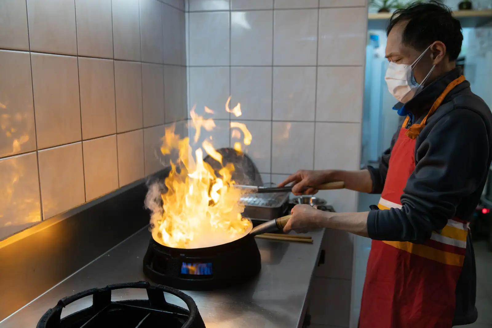 Chef cuisinant au wok, flamme vive, dans une cuisine professionnelle carrelée de restaurant asiatique.